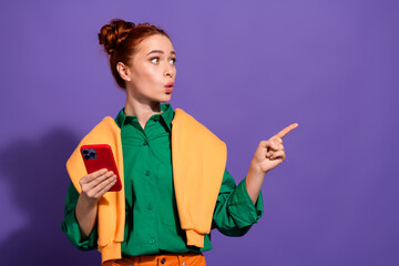 A stylish young woman with red hair gestures to the side while holding a smartphone, wearing a...