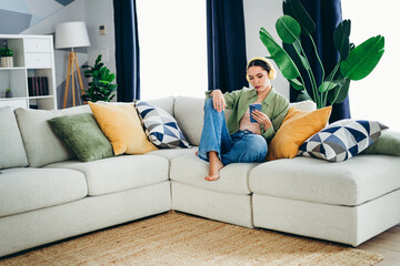 Obraz premium Charming woman relaxing on a stylish sofa with pillows and houseplants, enjoying a moment of leisure while using her smartphone