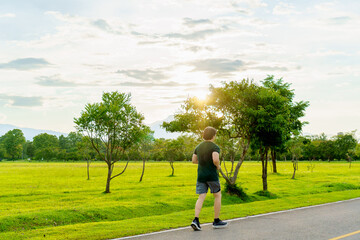 Sports asian man running on road for outdoor workout with warm light of sunset on freshy green of nature  background in rainy season. Jogging in countryside. Healthy lifestyle.