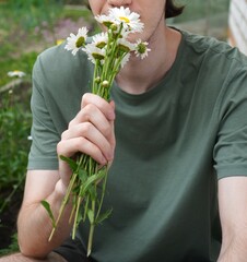  happy young farmer holds out a bouquet of daisy flowers in his hands, white summer flowers in his hair, sits on a stool near a garden bed with different flowering plants