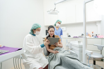 Obraz premium asian female dentist in protective gown and mask explains dental x-ray to asian adult female patient using tablet while assistant stands beside inside clean modern treatment room