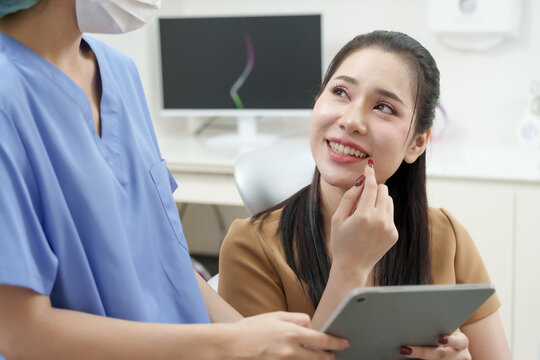 asian adult female patient points to lower tooth while smiling and talking with asian female dental assistant in blue uniform holding tablet during dental consultation inside bright clinic