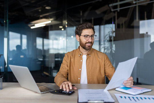 Successful businessman doing paperwork inside office. Male financier accountant checking tax reports, using calculator at workplace, satisfied with company results. - Powered by Adobe