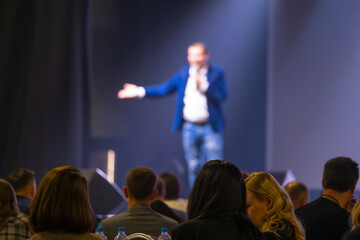 Conference setting with speaker delivering a presentation to audience in dim lighting.