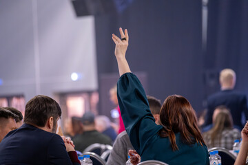 Person in crowded event raising hand to signal or ask a question. Back shot highlighting participation and engagement in a formal meeting or conference session.