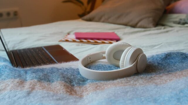 Close-up no people shot of white headphones, laptop, and pink notebook on bed in cozy teenage room