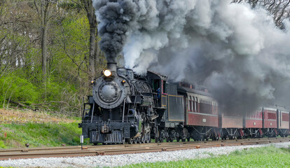 A vintage steam locomotive chugs along the tracks, billowing smoke into the air as it journeys through vibrant green fields. Spring foliage surrounds the scene, creating a picturesque setting.