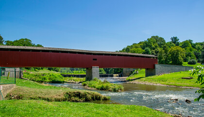 Obraz premium A red covered bridge crosses over a gentle stream surrounded by vibrant green trees on a bright, sunny day. Nature thrives in this peaceful rural setting, inviting exploration.