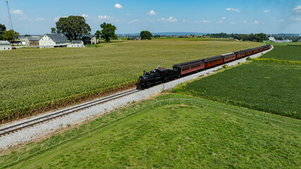 A vintage steam train navigates through lush green fields in Pennsylvania, surrounded by farmland. The clear blue sky above adds to the charm of this picturesque rural setting.