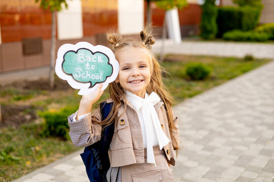 a first-grader girl with a sign in her hands back to school, a schoolgirl goes to school with a backpack or briefcase, a joyful child near school in September