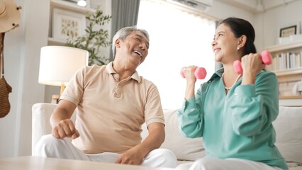 Fototapeta premium Happy elderly couple exercising together with dumbbells at home, focusing on muscle strength, joint health, and physical wellness for improved mobility and healthy aging. Retirement concept. Myrmidon.