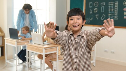 Asian boy looking at camera while waving hands and standing at blackboard. Skilled child greeting...