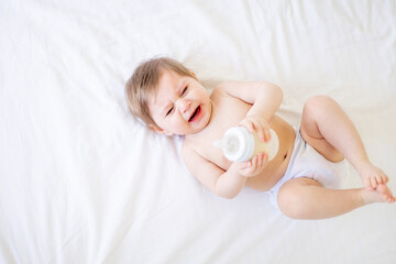 crying baby a newborn girl in a diaper lies on a white crib in a bright bedroom and sucks a...