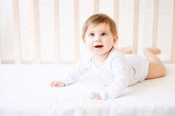 Fototapeta premium happy baby newborn girl on a white crib in a bright bedroom close-up in white clothes smiling or laughing, place for text, top view