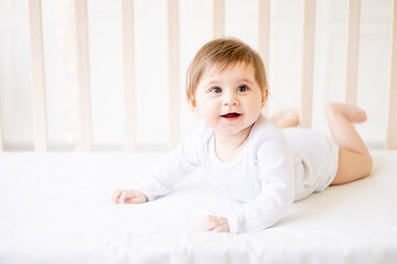 Fototapeta premium happy baby newborn girl on a white crib in a bright bedroom close-up in white clothes smiling or laughing, place for text, top view
