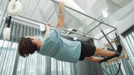 Man gripping bar on pilates trapeze table for core strength and upper body workout in a gym. Trainer hanging inverted back bend on the cadillac for improving flexibility in back and hip. Habituate.