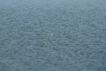 A bobber is seen floating on the water surface, gently swaying with the waves. The reflections and shadows create a serene setting in the mid-afternoon light