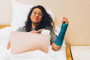 Woman with broken arm using laptop and smartphone in bed