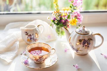 Creative postcard with porcelain cup of tea, teapot and a vibrant bouquet of garden flowers. Elegant tea set in beautiful soft sunlight. Good morning and teatime concept. Slow life visual trend.