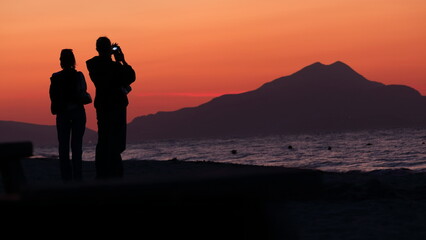 Sun sets over beach, people watching, peaceful glowing sea.