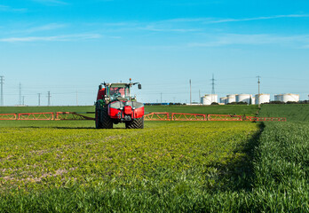 Fototapeta premium Tractor spraying crops in field with industrial tanks in the background
