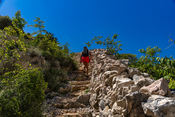 A woman in a white T-shirt and pink skirt walks along a hiking trail leading through the mountains from Baska to Vela Luka 