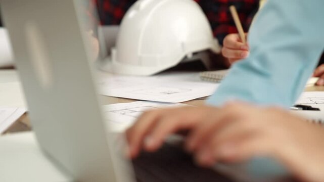 Architect and safety engineer discussion about building blueprint displaying on laptop on table with architectural document and wooden block scatter around at modern office. Closeup. Delineation.