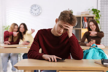 Cyber bullying. Upset teenage boy with smartphone and schoolmates pointing at him in classroom,...