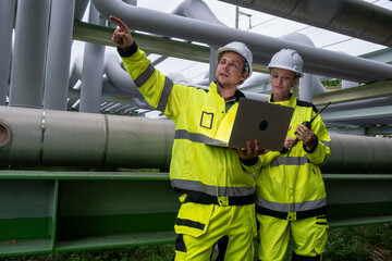Workers inspect industrial pipelines and manage operations at a processing facility during a sunny...