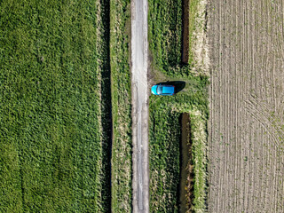 Bright blue car parked along a narrow dirt road surrounded by lush green fields on a sunny day in rural countryside