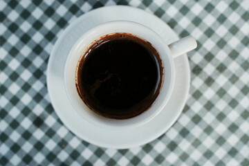 Top view of a cup of traditional Serbian coffee served on a white plate with checkered tablecloth.