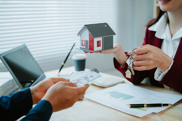 Asian female real estate agent in red shirt introduces house offer. Confident and friendly real estate agent presents small model home to large client.