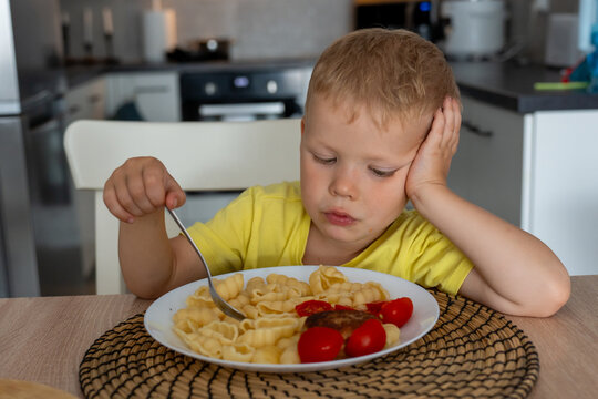 Bored little boy in yellow t-shirt sitting at kitchen table and staring at plate of pasta with no appetite. Concept of picky eating, childhood mood swings and mealtime struggles at home.