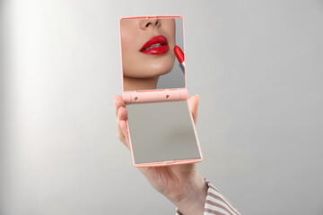 Beautiful woman looking in compact mirror while applying red lipstick on light grey background,...