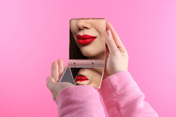 Beautiful woman with red lips looking in compact mirror on pink background, closeup © New Africa