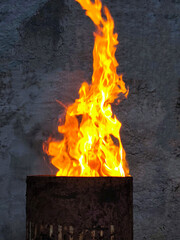 Bright flames rise from a rusty metal barrel against a rough concrete wall. Atmospheric photo of...