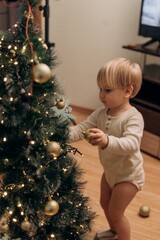 Little toddler carefully decorates Christmas tree with golden baubles at home capturing peaceful everyday moments of holiday preparation filled with wonder care learning and early independence