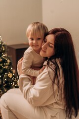 Mother cuddles child in soft pajamas beside festive tree evening light highlights intimate bond gentle warmth and quiet love between generations during peaceful seasonal moment at home