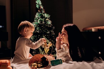 Smiling modern mother plays with son while decorating festive tree with golden ornaments sharing warmth and playful joy in a simple evening moment of holiday connection and gentle parenting