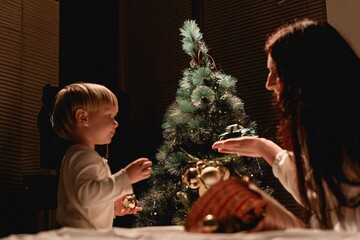 Mother and little boy bonding over decorating Christmas tree with baubles and toy truck during cozy evening filled with joy connection playful discovery and warm festive magic together