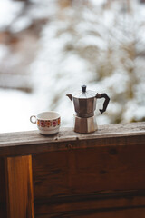 Moka pot and floral cup on wooden balcony with blurred snowy mountain landscape in the background. Peaceful winter coffee moment outdoors.