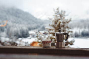Moka pot and floral cup on wooden balcony with blurred snowy mountain landscape in the background. Peaceful winter coffee moment outdoors.