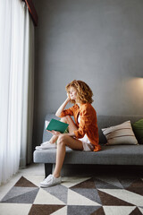 Young woman with curly hair sitting on a gray sofa, holding a green notebook, reflecting thoughtfully in a modern, minimalistic interior design