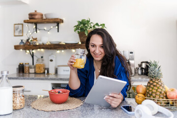 young latin woman overweight using tablet while having breakfast at home kitchen in Mexico Latin...