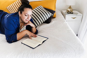 Young latin woman plus size reading book and lying on bed at bedroom in home in Mexico Latin...