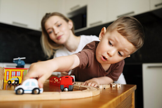 Little boy playing with excitement while pushing a toy car on a wooden train track, with his mother watching in the background at home.