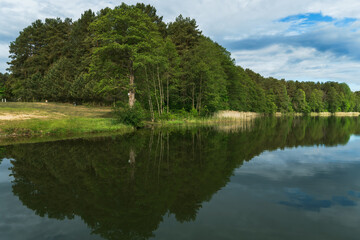 Sunny summer landscape on the lake. Reflection of the sky and clouds in the water. A valley of green trees on the shore of a lake. A concept for travel and camping.