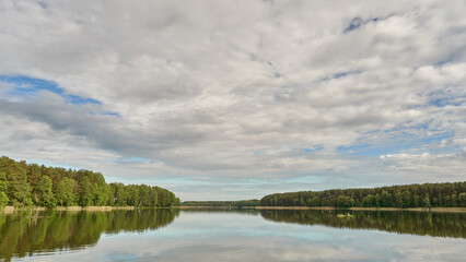 Sunny summer landscape on the lake. Reflection of the sky and clouds in the water. A valley of green trees on the shore of a lake. A concept for travel and camping.