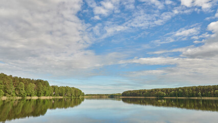 Sunny summer landscape on the lake. Reflection of the sky and clouds in the water. A valley of green trees on the shore of a lake. A concept for travel and camping.
