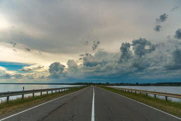 An empty asphalt road runs through the river. Thick gray clouds over the highway along the lake.